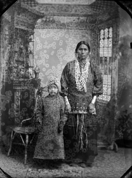 Studio full-length portrait of a Ho-Chunk girl posing standing on the left wearing a long winter coat and bonnet, and a Ho-Chunk woman posing standing on the right wearing several necklaces, a sash, traditional blouse, rings, earrings, file bracelets, and moccasins. Both are standing in front of a studio backdrop and have their hands at their sides.