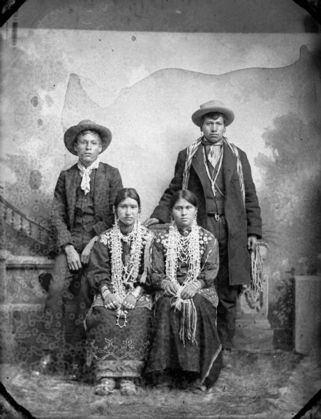 Studio portrait of two young Ho-Chunk women (unknown) seated, wearing several necklaces, earrings, traditional blouses, rings, and moccasins, in front of a prop stone wall. Two young men are behind them. On the left, George Monegar is sitting on the wall. He is wearing a suit coat, bandana, and hat, and Thomas Thunder (Hoonk Ha Ga Kah), standing on the right, is wearing an overcoat, earrings, a bandana, streamers, and a hat. In the background is a painted backdrop.