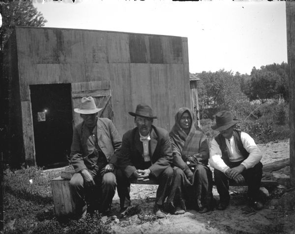 Three Ho-Chunk men wearing hats and a Ho-Chunk woman wrapped in a shawl are posing outdoors sitting on a bench in front of a corrugated metal building.