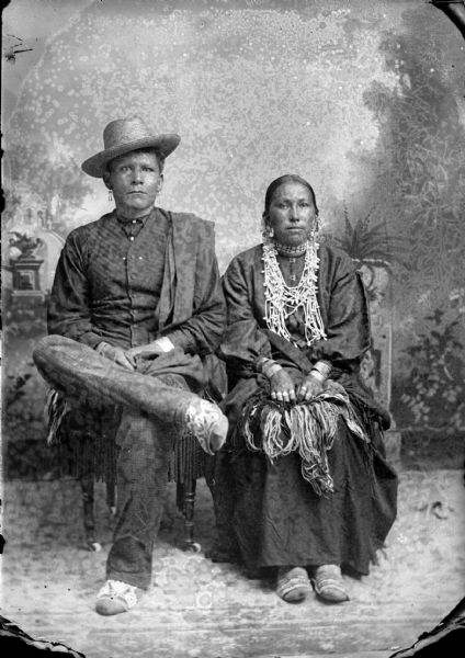 Studio portrait of a Ho-Chunk couple posing in front of a painted backdrop. The man sitting on the left is wearing a vest, straw hat, beaded moccasins, earrings, and a shawl over his left shoulder. A Ho-Chunk woman is sitting on the right wearing several necklaces, earrings, rings, a traditional blouse, and file bracelets.