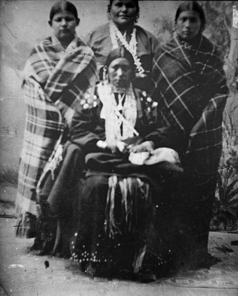 Copy photograph of a blurred studio portrait in front of a painted backdrop of a Ho-Chunk woman posing sitting in front of three other Ho-Chunk women posing standing. All of the women are wearing several necklaces and earrings, and the women standing on the outside are wrapped in plaid shawls. The top of the head of woman standing center is cut off.