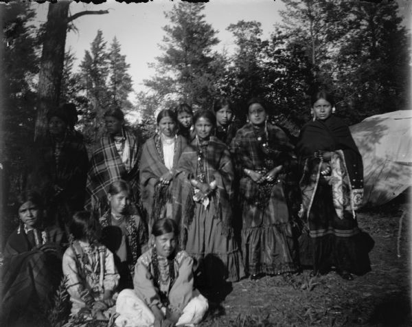 Outdoor portrait of twelve Ho-Chunk women and girls posing standing and sitting in front of several trees, possibly at a powwow. They are all wearing many necklaces and earrings, and many have shawls wrapped around their shoulders. In the background on the right side is a structure, possibly a type of lodge.