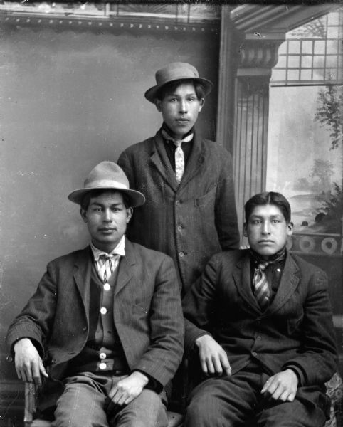 Studio portrait of three Ho-Chunk men posing in front of a painted backdrop. A Ho-Chunk man is sitting on the left wearing a suit coat, button-down sweater, and hat. Another Ho-Chunk man is sitting on the right wearing a suit coat and a necktie. The third man is standing behind them wearing a suit coat, necktie, and hat.