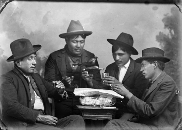 Studio portrait of four Ho-Chunk men posing sitting in front of a painted backdrop. They are sitting around a table on which is sitting two packages wrapped in newspapers, and a clay pipe. The men are wearing suit coats and hats. The men sitting on the far left and right are holding small glasses, and the man third from the left is pouring from a liquor bottle into a small glass. Above the liquor bottle is a stuffed bird, possibly a crow. The man second from the left is holding a clay pipe. They are identified as members of the Greencrow family.