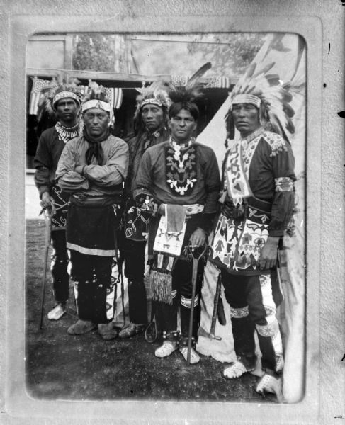 Copy photograph of five Ho-Chunk men posing standing outside tents, possibly at a powwow. They are dressed in full regalia, including headdresses, bandoleers, and moccasins.