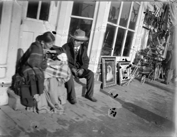 A Ho-Chunk man, woman, and child are sitting on steps in front of a storefront on a board sidewalk. Along the sidewalk in front of windows are two paintings and stacks of wagons and chairs suspended from the building near an awning. On the far right another man is standing near the stack of chairs.