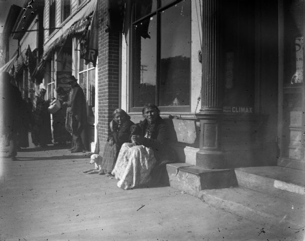 Two elderly Ho-Chunk women posing sitting on a storefront curb with two dogs. Probably in front of the Jones Lumber and Mercantile Store on Water Street. There are other people standing on the sidewalk near a doorway on the left.