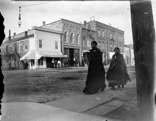 Two Ho-Chunk women walking on the board sidewalk on Main Street at the intersection of Main and Second Streets. They are both wearing shawls wrapped around their shoulders. Pedestrians walk on the sidewalk in front of buildings across the street. An awning in front of a buildings reads: Kelley's Confectionery."