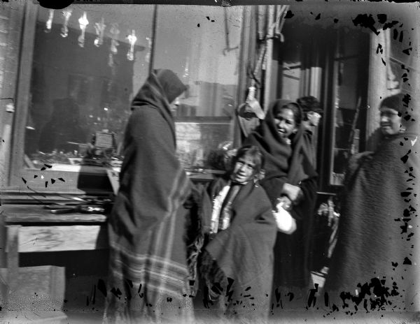 Three Ho-Chunk women and two children are standing in front of a storefront. The Ho-Chunk women and girl are all wearing shawls wrapped around their heads and shoulders. The boy near the entrance is wearing a cap. Behind them is a shop window.