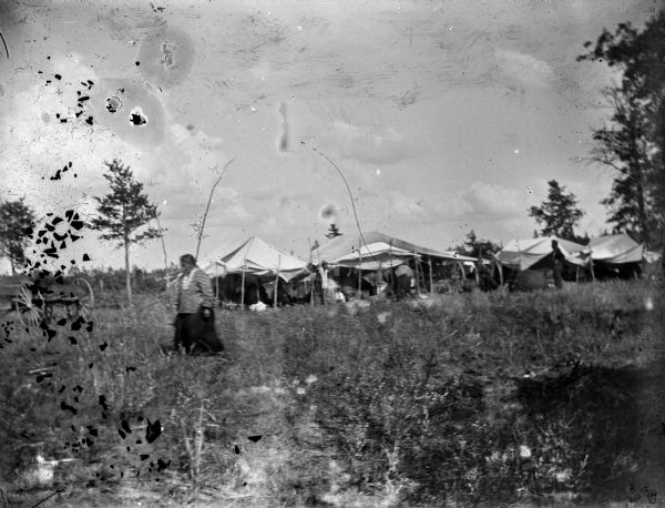 A Ho-Chunk woman walking in a field. In the background are several white tents, possibly an encampment for a cranberry harvest.