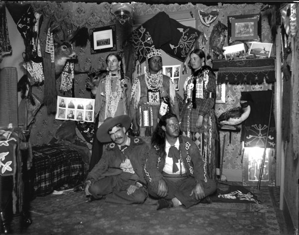 Two European American men posing sitting in front of two European American women and a Ho-Chunk woman posing standing. They are all wearing Ho-Chunk regalia in a room decorated with Ho-Chunk artifacts and photographs. The man sitting on the left is Tom Roddy.