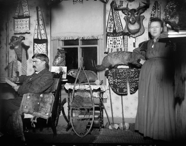 A European American man is posing reading on the left, and a European American woman is posing standing on the right in a room decorated with Ho-Chunk artifacts, stuffed animals, and sporting equipment. They are identified as Tom Roddy and his wife at home.