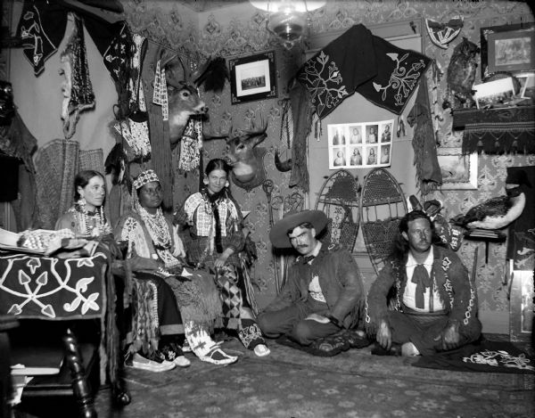 Two European American men are posing sitting on the floor cross-legged and kneeling on the right, and two European American women and a Ho-Chunk woman posing sitting on the left. They are all wearing Ho-Chunk regalia in a room decorated with Ho-Chunk artifacts and photographs. The man sitting second from the right is Tom Roddy, and the artifacts are from the collection of Tom Roddy.
