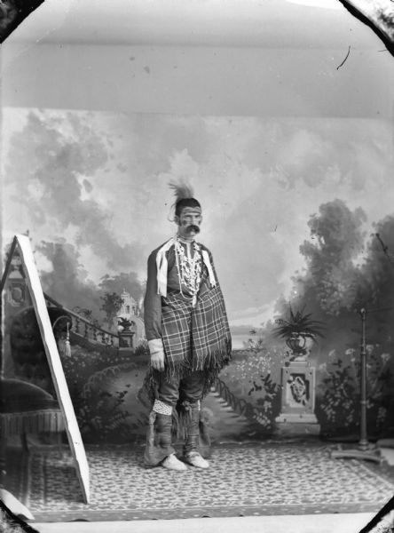 A studio portrait of Frank Long, a European American man with a moustache posing in front of a painted backdrop before going to a costume party. He is wearing a Ho-Chunk costume, which includes a feather in his hair, war paint, garters, shirt, necklaces, moccasins, and a shawl wrapped around his torso.
