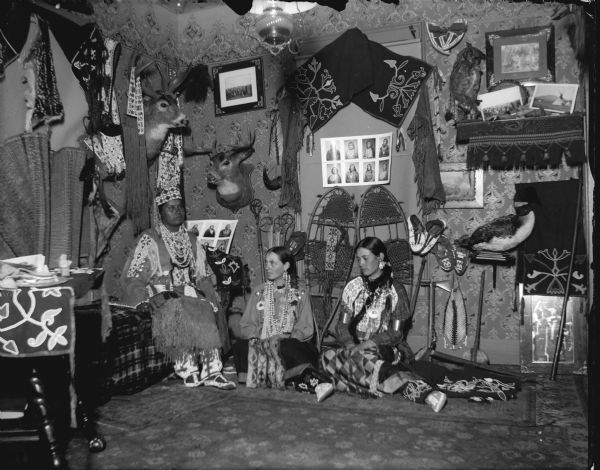 Group portrait of a Ho-Chunk woman posing sitting on a bed or sofa, and two European American women sitting on the floor, all dressed in Ho-Chunk regalia. They are in a room decorated with Ho-Chunk artifacts and photographs. The artifacts are probably from the collection of Tom Roddy.