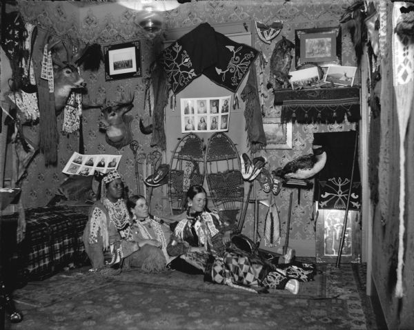 Two European American women posing reclining against a Ho-Chunk woman posing sitting on the floor against a couch. They are dressed in Ho-Chunk regalia. The room is decorated with Ho-Chunk artifacts and photographs, and the artifacts are probably from the collection of Tom Roddy.