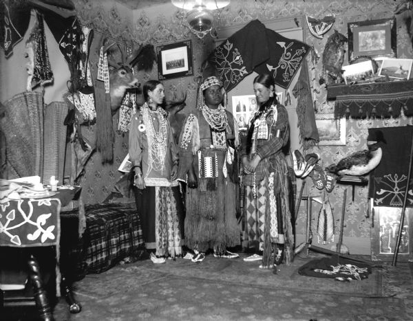 A portrait of a Ho-Chunk woman posing standing between two European American women also posing standing. All are wearing Ho-Chunk regalia, and are in a room decorated with Ho-Chunk artifacts and photographs. The artifacts are probably from the collection of Tom Roddy.