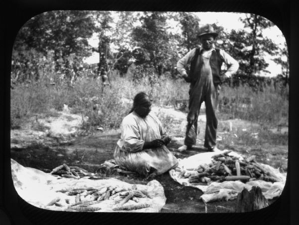 Lantern slide of a Ho-Chunk woman posing sitting on the ground in a clearing with trees in the background. She is sitting cross-legged and is holding an ear of corn near two cloth sheets full of corn. A Ho-Chunk man is posing standing on the right, with his hands on his hips and wearing a hat. They are identified as Joe Bear Heart and his wife.