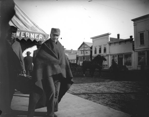 A Ho-Chunk man wearing a cap and wrapped in a shawl is walking near the front of the Werner Drugstore on Main Street between Water and First Streets. There are other people in the background, and two horses pulling a carriage in the street.