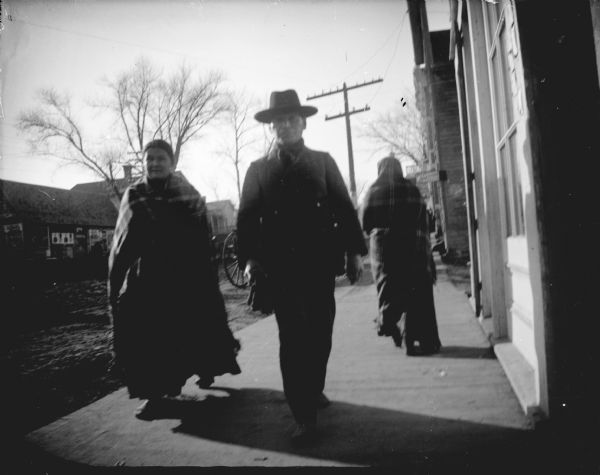 A Ho-Chunk man and two Ho-Chunk women are walking on the north side of Main Street between Second and Third Street. The man is wearing a hat and the woman is wearing a shawl wrapped around her shoulders.