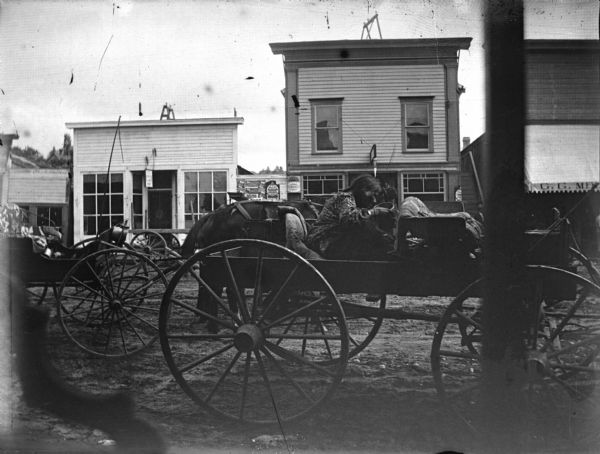 A Ho-Chunk woman sitting in a wagon on the north side of Main Street between Second and Third Streets. In the background there are several other wagons, a horse, and storefronts across the street.