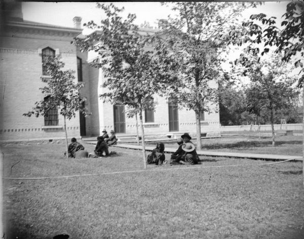 Ho-Chunk men and women sitting outside on the lawn of a large building with bars. Identified as probably the family of Jim Swallow at the County Jail. Jim Swallow was in jail for killing George Black Hawk in 1895.