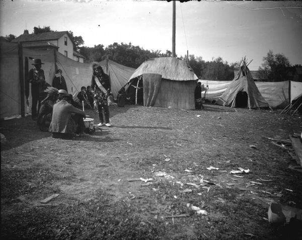 Ho-Chunk men are sitting and standing in an area portioned off by canvas barriers and a lodge and teepee. Identified as probably the Homecoming Powwow in 1908 at the intersection of Main and Second Streets. Dwellings can be seen in the background on a hill.
