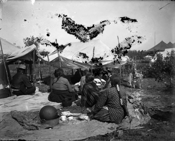 Ho-Chunk women and men sitting in front of and under cloth awnings on blankets with their backs to the camera, probably a powwow. On the blankets are also cooking utensils and dishes. Large tents are in the background on the right.