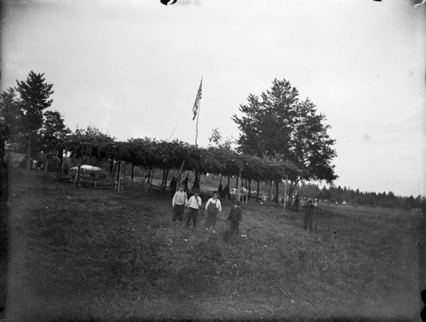 Four Ho-Chunk children and one more off to the right are posing standing in front of an arbor with an United States flag on a long pole, probably at a powwow. Other people, tents, and trees are in the background.