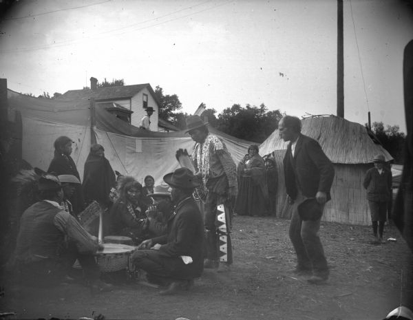 Ho-Chunk men and women sitting and standing around a large medicine drum in an area portioned off by canvas barriers in front of a lodge. Identified as the Homecoming Powwow in 1908 at the intersection of Main and Second Streets. A house on a hill is in the background.