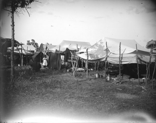 Ho-Chunk awnings and tents with a few individuals nearby, including a child in a light-colored dress, and a woman with a long braid sitting in the grass. Probably a powwow encampment.