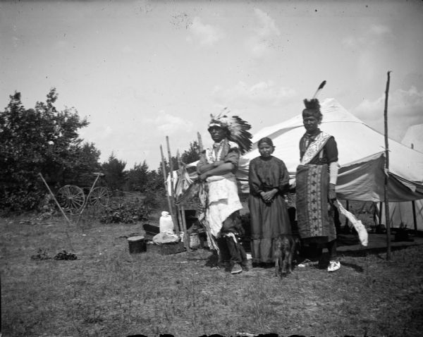 A Ho-Chunk woman, standing between two Ho-Chunk men dressed in regalia, including feathered headdresses, in front of an awning. Probably at a powwow. A dog is walking between the woman and man on the right.