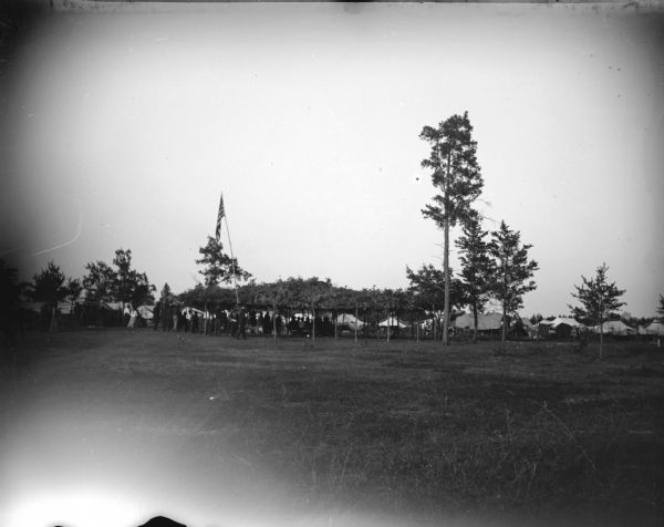 View from distance of an arbor, with the United States flag on a long pole and individuals underneath it. Tents are in the background. Probably at a Ho-Chunk powwow.