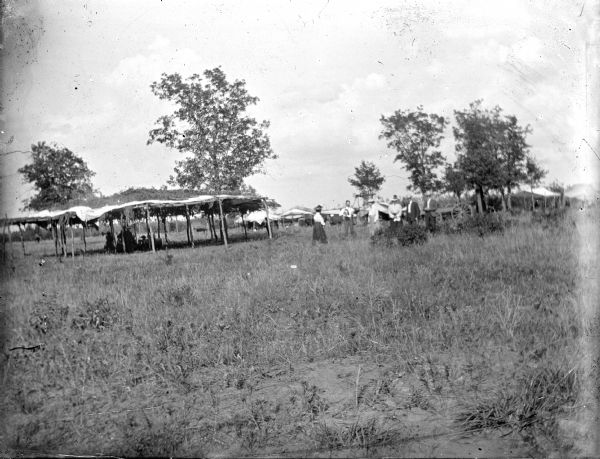 View from a distance towards an arbor, with tents and a few awnings surrounding it. A few individuals are sitting underneath in the shade. Possibly a Ho-Chunk powwow with a dance ring. A small group of European Americans, including men and women, are standing on the right in the long grass.