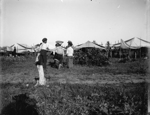 View towards children wearing caps and standing in a field, with tents and awnings in the distance. Possibly a Ho-Chunk powwow encampment. The shadow of a man is in the foreground; perhaps the photographer.