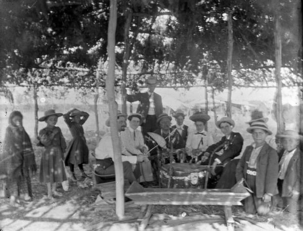 European American men, women, and children posing sitting and standing underneath an arbor around a medicine drum and a Ho-Chunk man.