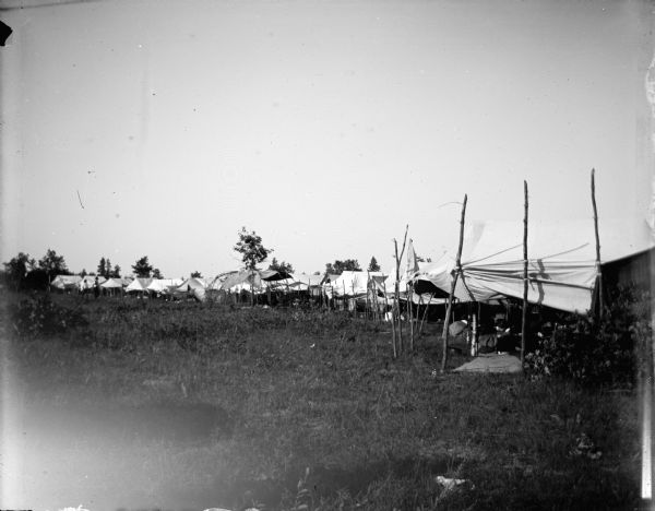 View from distance of several awnings and tents lining a field with several individuals. Possibly a Ho-Chunk powwow encampment.