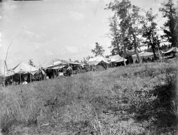 View from distance of several awnings lining a field. Individuals can be seen around and underneath them. Possibly a Ho-Chunk powwow encampment.