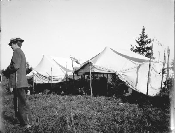 A European American man holding a cane and wearing a suit and straw hat is standing on the left and looking left. He is in front of an awning in a field. Individuals are underneath the awnings. Possibly a Ho-Chunk powwow encampment.