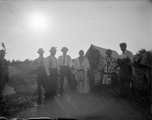 Ho-Chunk men, women, and child posing standing in front of a tent. The four men are wearing hats, and the three women are wearing more traditional dress. The child is standing in the center near the fourth man from the left.