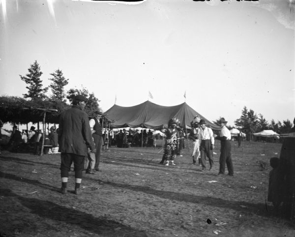Ho-Chunk and European American men and boys in front of an arbor and tent with United States flags on top. Probably meeting of the Blue Wing Band at Valley Junction on the Wyatt and Purdy Meadow.