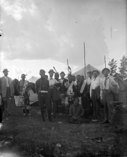 Several Ho-Chunk men, women, and children, and a few European American men, posing standing in front of tents in a field. They are members of the Blue Wing Band at Valley Junction on the Wyatt and Purdy Meadow.