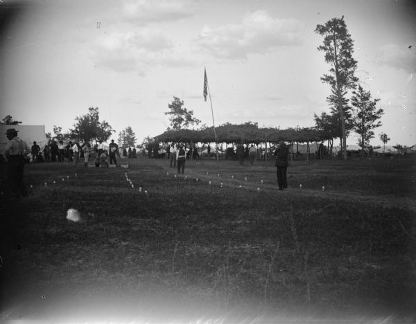 View from field of people standing near lines of glass bottles in the grass. In the background are tents, and an arbor and several individuals underneath, and the United States flag on a pole in front. Several European American men are in front of the arbor.