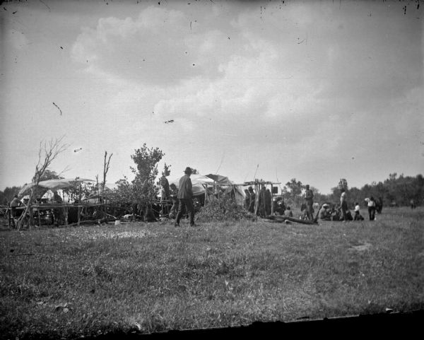 View from field of several individuals around a large pile of brush under spits near a rough short fence and lodge. Possibly a feast fire at a powwow.