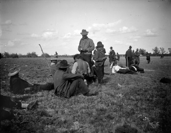 Ho-Chunk men and boys sitting and standing in a field, probably watching a baseball game.