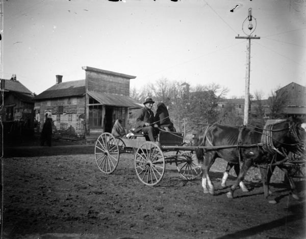 Ho-Chunk man and two Ho-Chunk women in a wagon pulled by a two-horse team. They are probably about to cross the bridge toward the Mission at the intersection of Water and Harrison Streets. There are buildings, power lines, and what looks like a church building in the background.