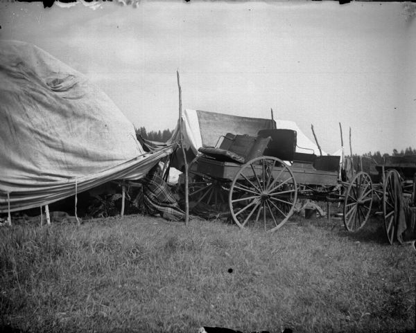 Ho-Chunk women and children sitting underneath white tents next to parked wagons.