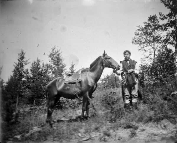 A Ho-Chunk man posing standing on a wooded hillside displaying a single horse with a saddle.