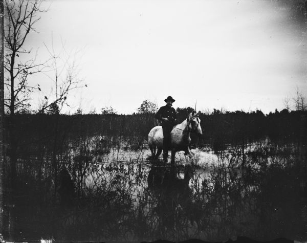 A man wearing a hat, possibly Ho-Chunk, is mounted bareback on a horse knee-deep in water in a marsh.