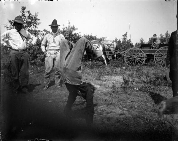 Ho-Chunk man performing a handstand in front of two Ho-Chunk men standing on the left. There is a child sitting in one of several horses horse-drawn wagons in the background.